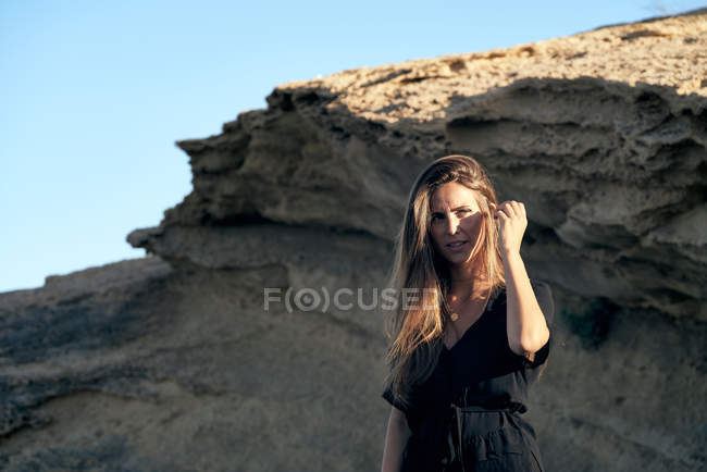 Junge langhaarige stilvolle nachdenkliche Frau steht im Sonnenlicht mit Felsen auf dem Hintergrund — Stockfoto
