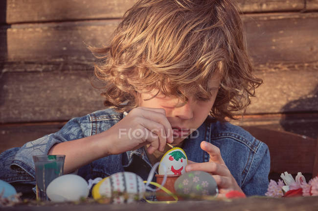 Adorable enfant assis et peignant des œufs avec une couleur vive à la table en bois — Photo de stock