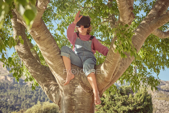 Энтузиастичный мальчик в очках виртуальной реальности и джинсовом комбинезоне сидит высоко на дереве в яркий день — стоковое фото