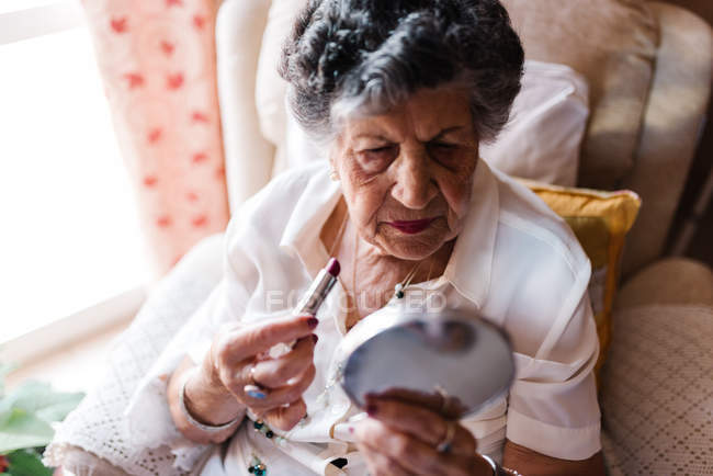 Пожилая женщина наносит помаду, глядя в зеркало и сидя на кресле дома — стоковое фото