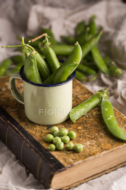 Fresh peas and pea pods in enamel cup on old shabby book — Stock Photo