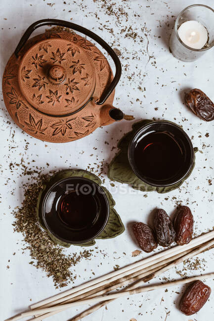 Зверху ароматний смачний чай у чашці з глиняним чайником та солодкими фініками на білому підносі, прикрашеному чайним листям на дерев'яному фоні — стокове фото