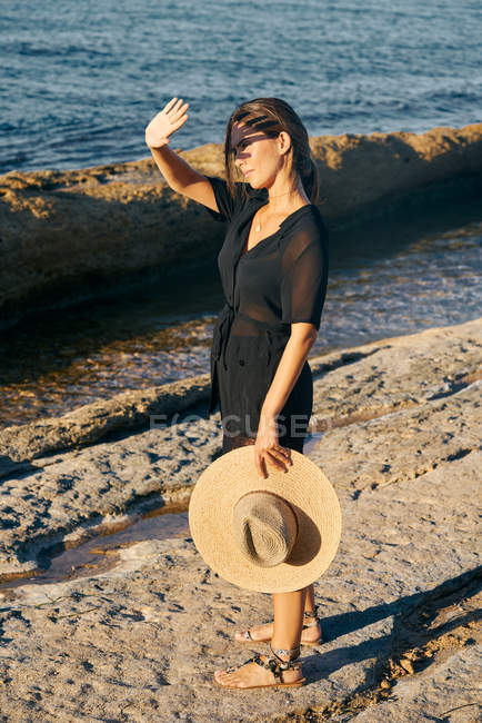 Giovane donna attraente che copre il viso dal sole sulla spiaggia e tenendo il cappello — Foto stock