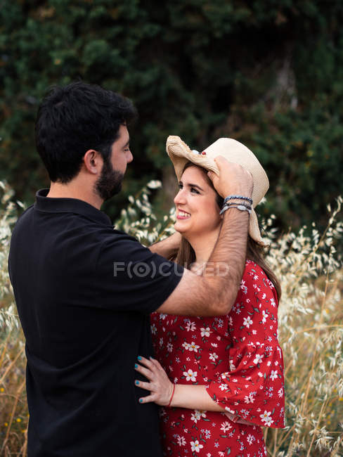 Man hugging smiling pregnant wife while adjusting straw hat on background of picturesque green park — Stock Photo