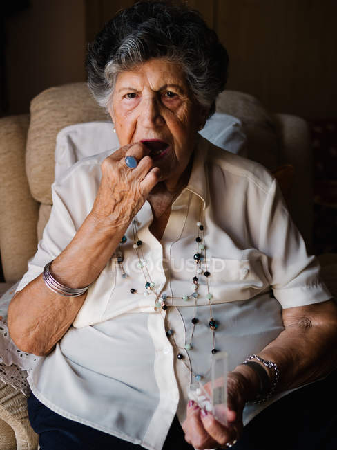 Elegante mujer mayor tomando pastillas, sentado en el sillón en casa y mirando a la cámara - foto de stock