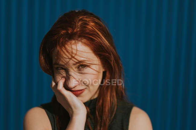 Mujer sonriente entrecerrando los ojos a la luz del sol y disfrutando del clima contra la pared azul - foto de stock