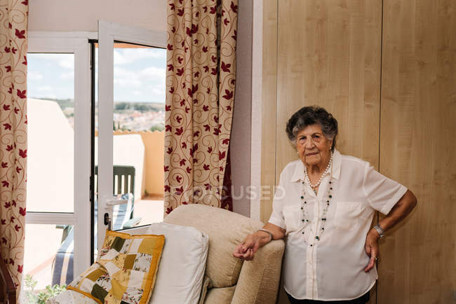 Seniorin im weißen Hemd mit der Hand auf der Taille steht, während sie sich zu Hause auf einen Sessel lehnt und in die Kamera schaut — Stockfoto