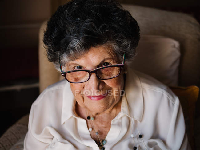 Retrato de mujer mayor feliz en camisa blanca y con cuentas en el cuello mirando a la cámara en casa - foto de stock