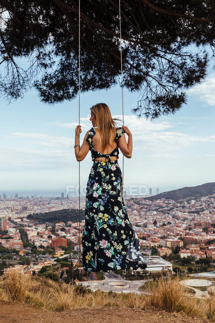 Mädchen auf dem Rücken in einem Blumenkleid, das die Stadt betrachtet, während sie auf einer Schaukel klettert — Stockfoto