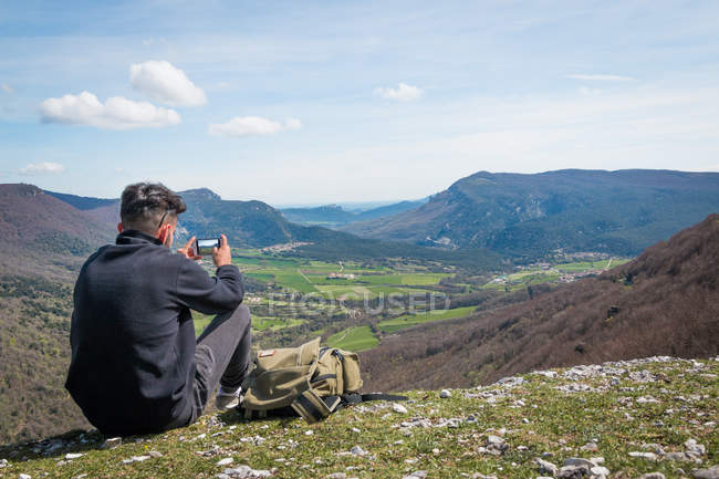 Вид ззаду чоловічого мандрівника, що сидить на горі і фотографує мальовничий вид на горбисту долину за допомогою мобільного телефону — стокове фото