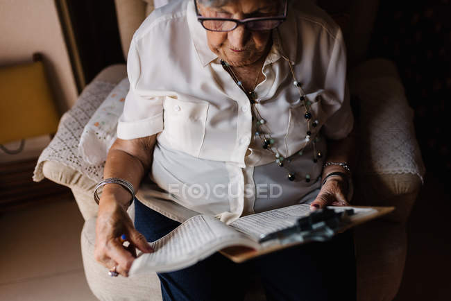 Donna anziana in camicetta e occhiali seduta sulla poltrona vicino alla finestra e risolvere cruciverba a casa — Foto stock
