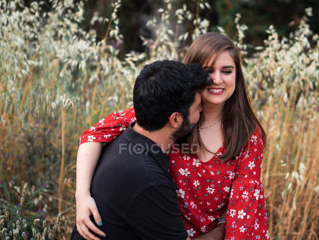 Чоловік обіймає усміхнену вагітну дружину, сидячи на колінах на фоні мальовничого зеленого парку в сонячний день — стокове фото