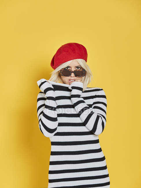 Cheerful blonde woman in sunglasses and striped blouse and red French cap smiling on yellow background — Stock Photo