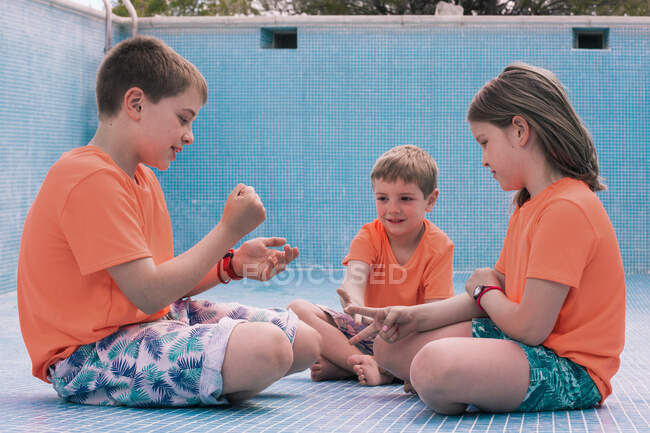 Enfants en T-shirts orange vif assis sur le fond de la piscine vide et jouant pierre-papier-ciseaux — Photo de stock