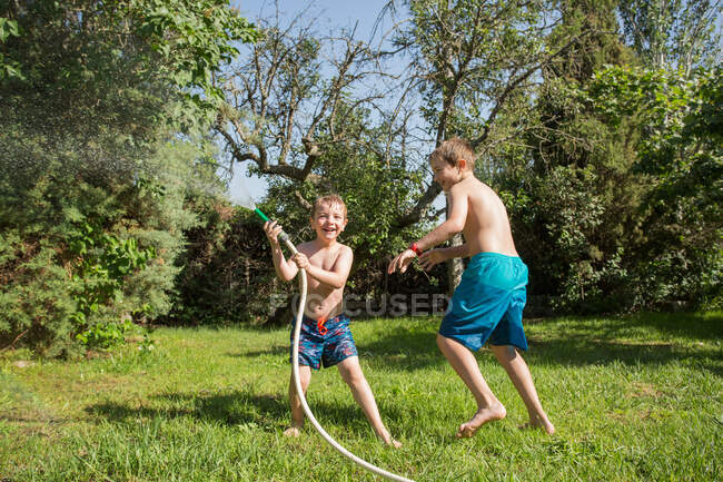 Kleine Kinder in Badebekleidung rennen herum und spritzen sich gegenseitig Wasser aus Gartenschlauch zu — Stockfoto