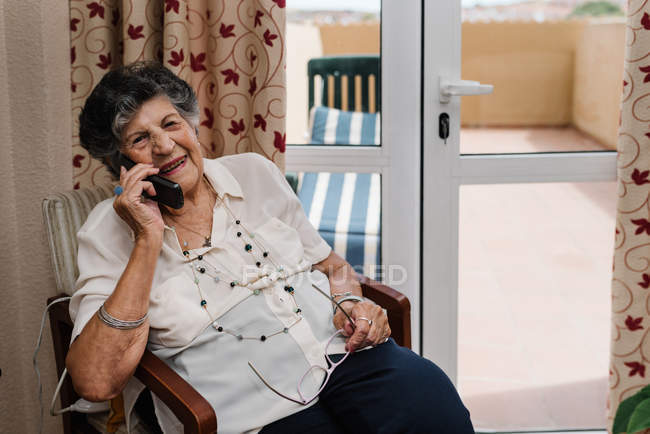 Feliz mujer mayor sonriendo y hablando en el teléfono móvil mientras mira hacia fuera en casa - foto de stock