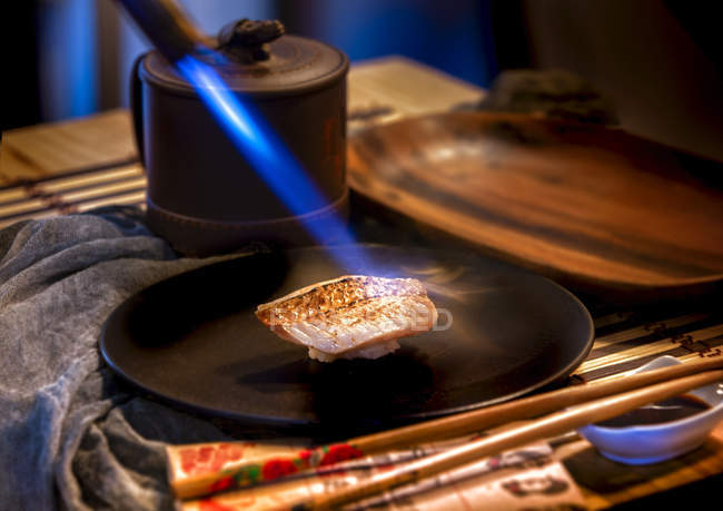 Голубой свет горелки готовит кусок рыбы на специальной тарелке и соус с палочками на столе — стоковое фото
