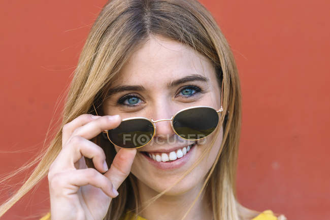 Портрет усміхненої молодої жінки з блакитними очима знімає сонцезахисні окуляри і посміхається на камеру — стокове фото