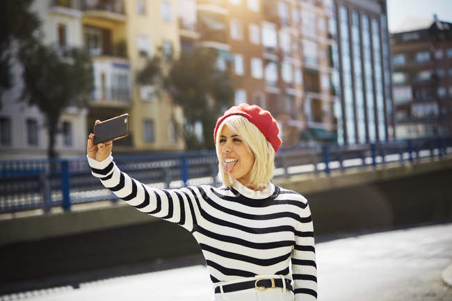 Jeune femme souriante en bonnet rouge français, chemisier rayé et short blanc prenant des photos sur fond urbain — Photo de stock