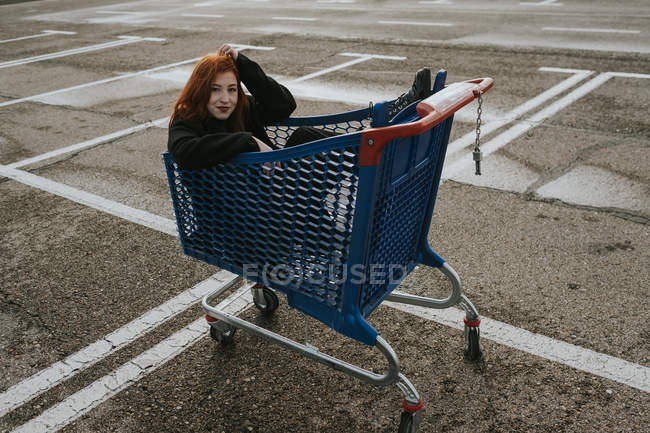 Atractiva joven con el pelo rojo en chaqueta negra divirtiéndose sentada en el carrito de la compra en el estacionamiento marcado - foto de stock
