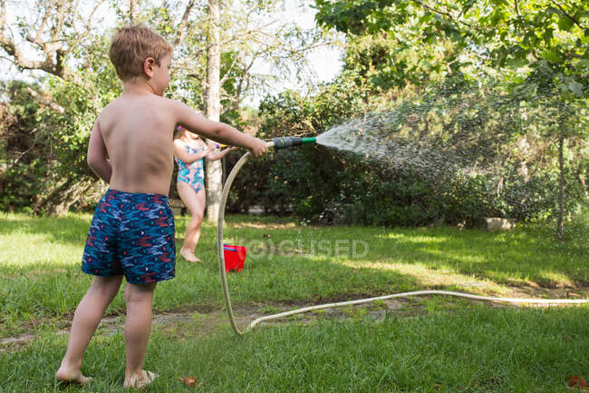 I bambini piccoli in costume da bagno che corrono in giro e spruzzano acqua dal tubo da giardino l'uno contro l'altro — Foto stock