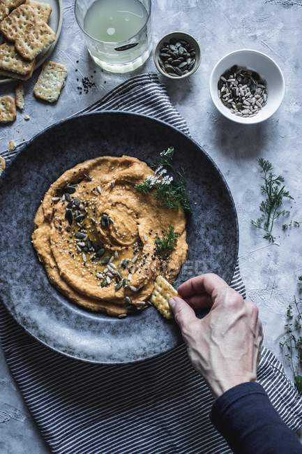 From above hand of person dunking cracker in plate with carrot and chickpea hummus with seeds — Stock Photo
