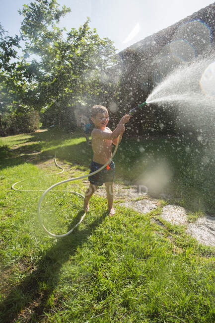 Маленький смеющийся ребенок в шортах и с босыми ногами брызгает водой из садового шланга — стоковое фото