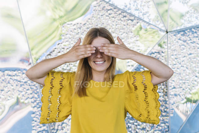 Joven mujer juguetona cubriendo los ojos con las manos sobre fondo metálico - foto de stock