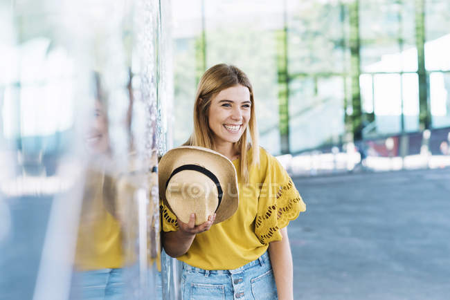 Young blonde woman leaning on wall and holding a straw hat — Stock Photo