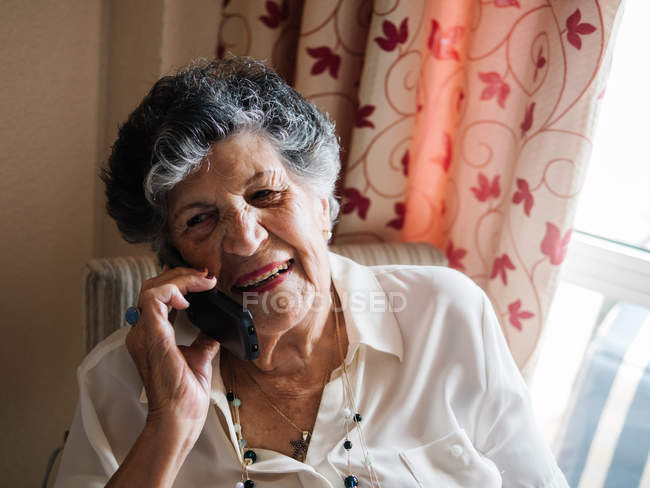 Mulher sênior feliz sorrindo e falando no telefone celular enquanto olha para longe em casa — Fotografia de Stock