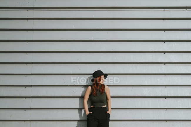 Femme en chapeau noir appuyé sur un mur gris rayé — Photo de stock