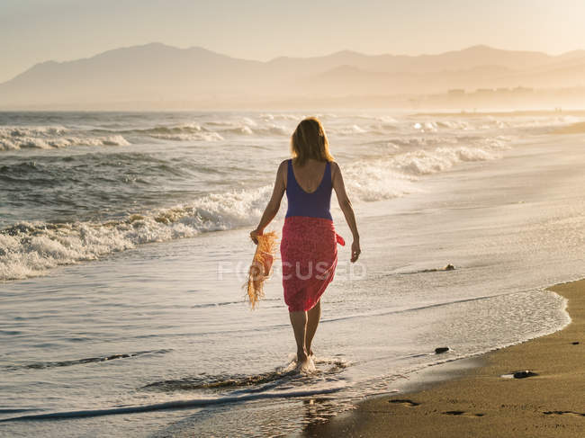 Back view of slim woman with hat and pareo walking along coastline in bright sunlight — Stock Photo