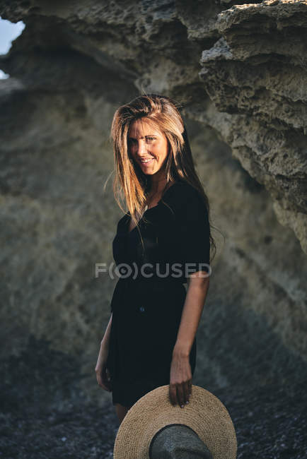 Giovane donna dai capelli lunghi sorridente guardando la fotocamera che tiene il cappello davanti alla roccia — Foto stock