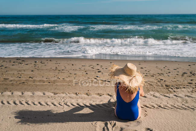Rückansicht einer hübschen Frau mit Hut und Badeanzug, die am Sandstrand sitzt und die Wellen unter türkisfarbenem wolkenverhangenem Himmel betrachtet — Stockfoto