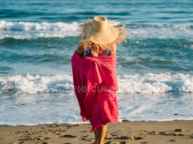 Unrecognizable woman in hat wrapped in pink scarf and walking along seaside in sunny bright day — Stock Photo