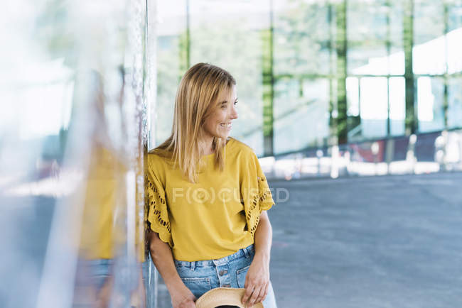 Young blonde woman leaning on wall and looking away, holding a straw hat — Stock Photo