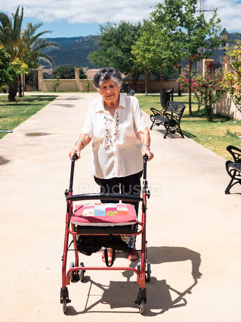 Glückliche Seniorin in Bluse mit Rollator an sonnigem Tag im tropischen Park — Stockfoto