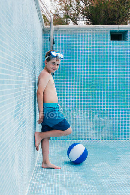 Garoto masculino em óculos com bola ao lado da parede da piscina vazia e olhando para a câmera — Fotografia de Stock