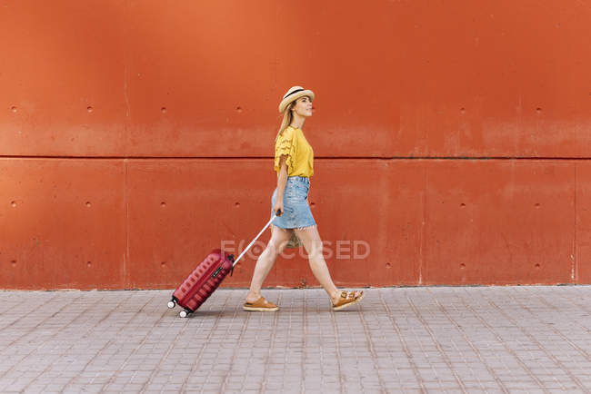 Young female tourist walking with suitcase against red wall on street — Stock Photo