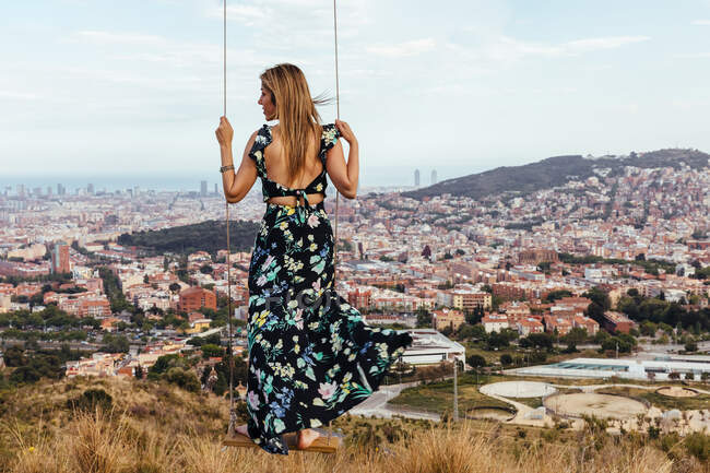 Fille sur le dos dans une robe florale contemplant la ville tout en grimpant sur une balançoire — Photo de stock