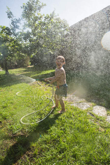 Маленький смеющийся ребенок в шортах и с босыми ногами брызгает водой из садового шланга — стоковое фото