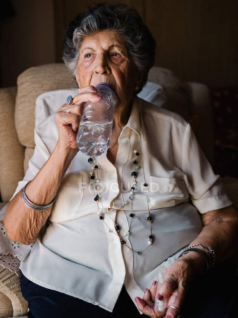 Старша жінка в білих пігулках п'є пігулки з водою з пляшки, сидить на кріслі і озирається в квартирі — стокове фото