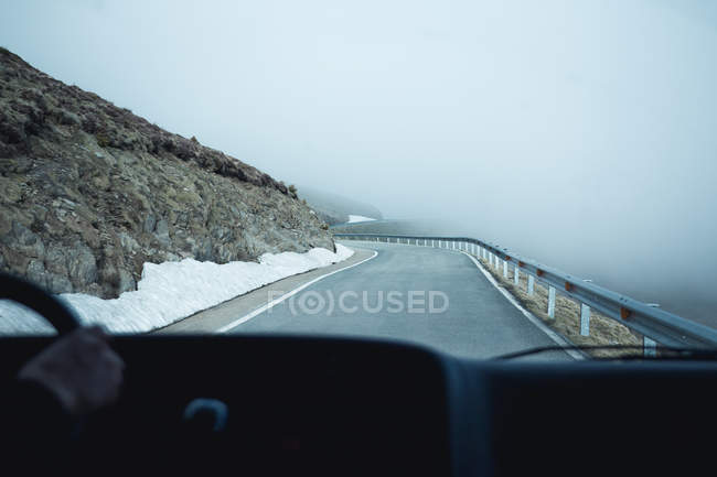 Hand of anonymous person driving car on asphalt road through snowy mountainous terrain on misty day — Stock Photo