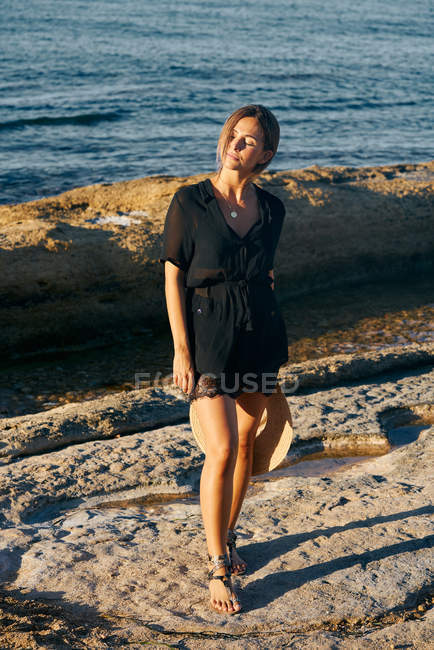 Mujer atractiva joven disfrutando del sol con los ojos cerrados en la playa y sosteniendo el sombrero - foto de stock
