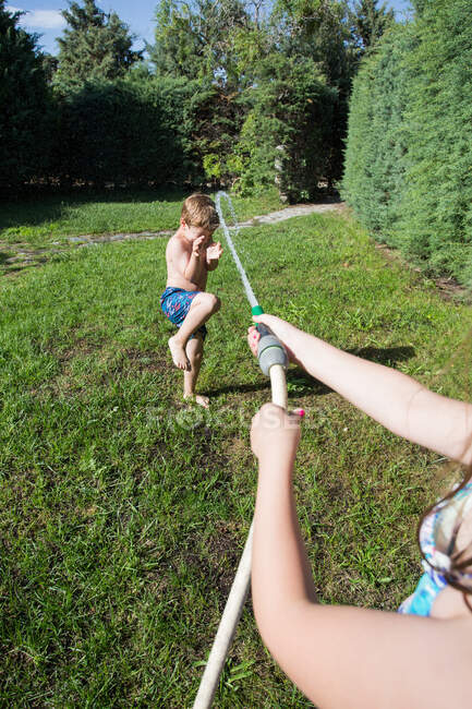 Bambini piccoli in costume da bagno che corrono in giro e spruzzano acqua dal tubo da giardino l'uno contro l'altro, vista in prima persona — Foto stock
