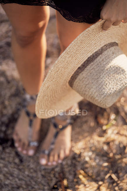 Primer plano de la mujer de pie y sosteniendo el sombrero en la mano en las sandalias de verano - foto de stock