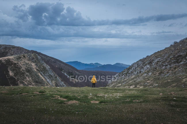 Back view of tourist in yellow coat looking at rough hills and overcast sky during trip in countryside — Stock Photo