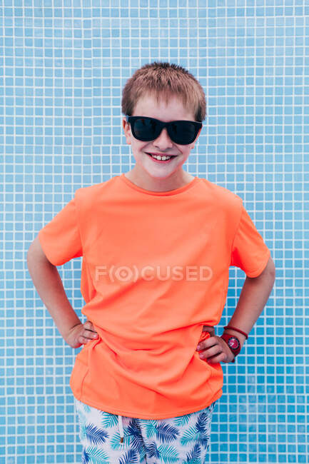 Enfant mâle souriant en T-shirt orange vif et lunettes de soleil debout sur fond de mur bleu carrelé — Photo de stock