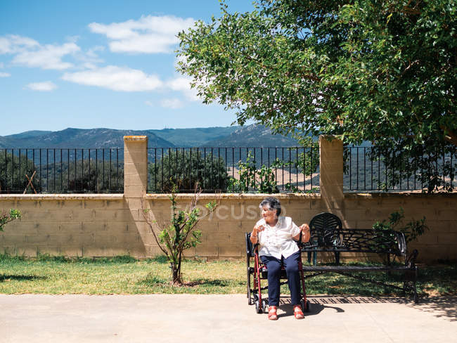 Senior woman in blouse sitting in walker in tropical park on sunny day — Stock Photo