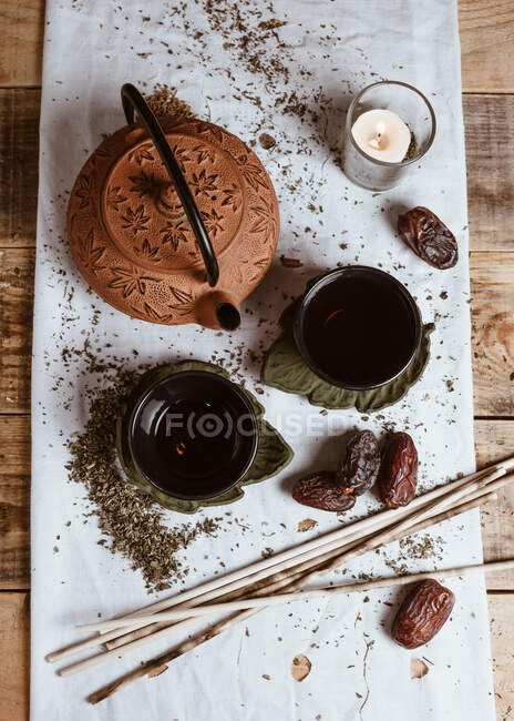 Зверху ароматний смачний чай у чашці з глиняним чайником та солодкими фініками на білому підносі, прикрашеному чайним листям на дерев'яному фоні — стокове фото